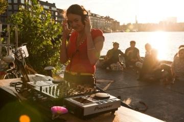 Vrouw staat achter een draaitafel muziek te maken. Achter haar zitten jongeren te chillen in de zon.