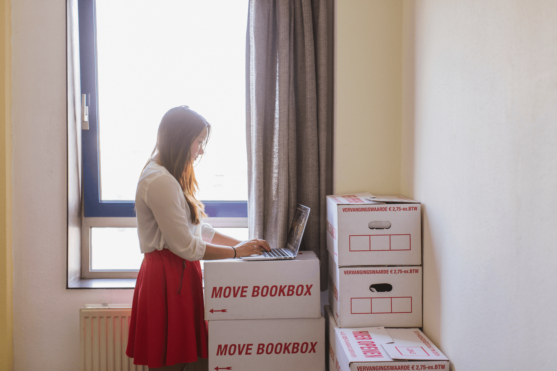 Een vrouw naast opgestapelde verhuisdozen met een laptop op de dozen. Ze zoekt naar een nieuwe woning.