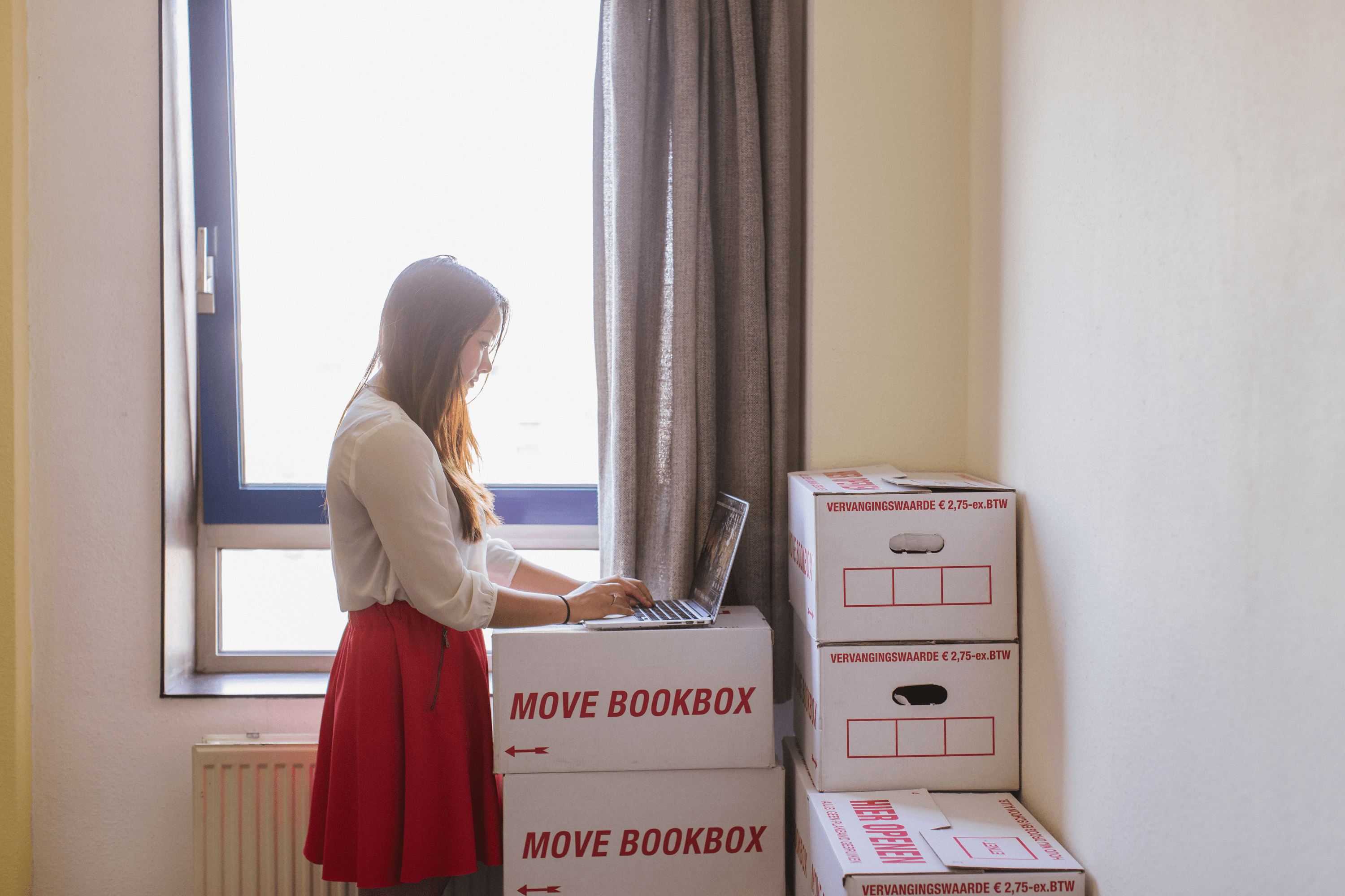 Een vrouw naast opgestapelde verhuisdozen met een laptop op de dozen. Ze zoekt naar een nieuwe woning.