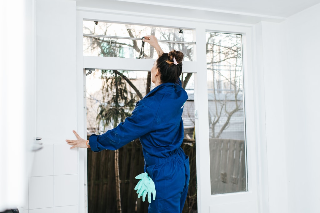 Een vrouw in een blauwe overall en schoonmaakhandschoenen maakt een raam open.